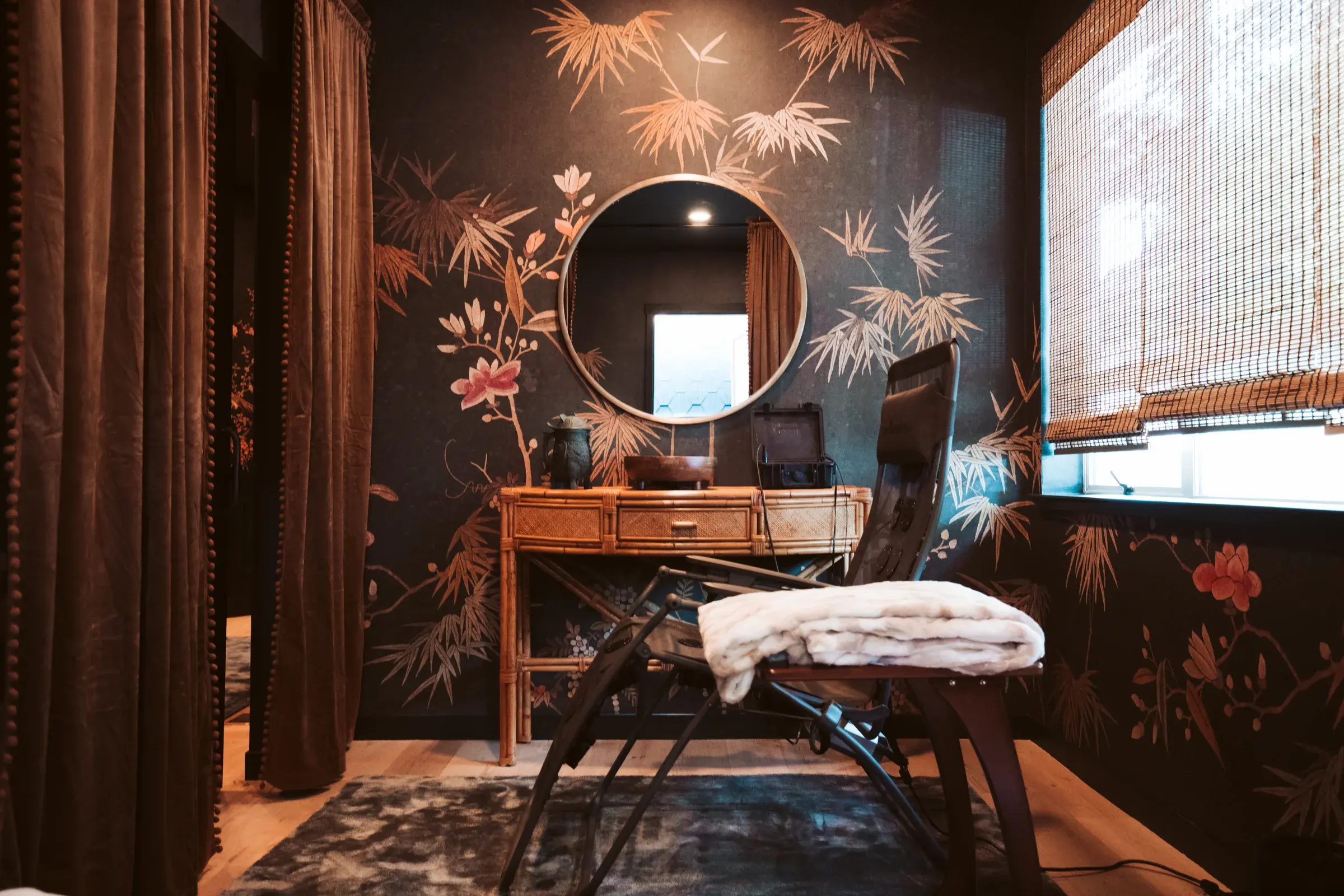 Treatment room with reclining lounger, fur throw, rattan desk, round mirror and dark floral mural wall