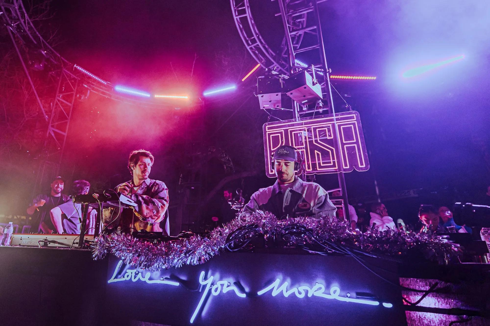 Two DJs under a neon RASA marquee with a roller coaster loop overhead, pink and blue stage light