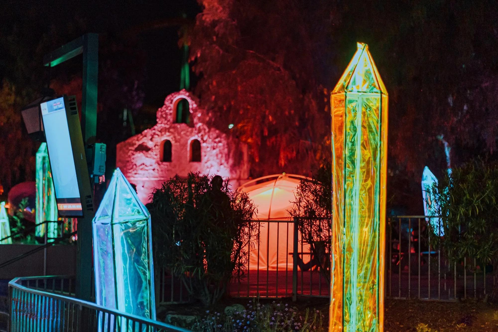 Glowing neon obelisk sculptures beside a red-lit mission facade under night trees