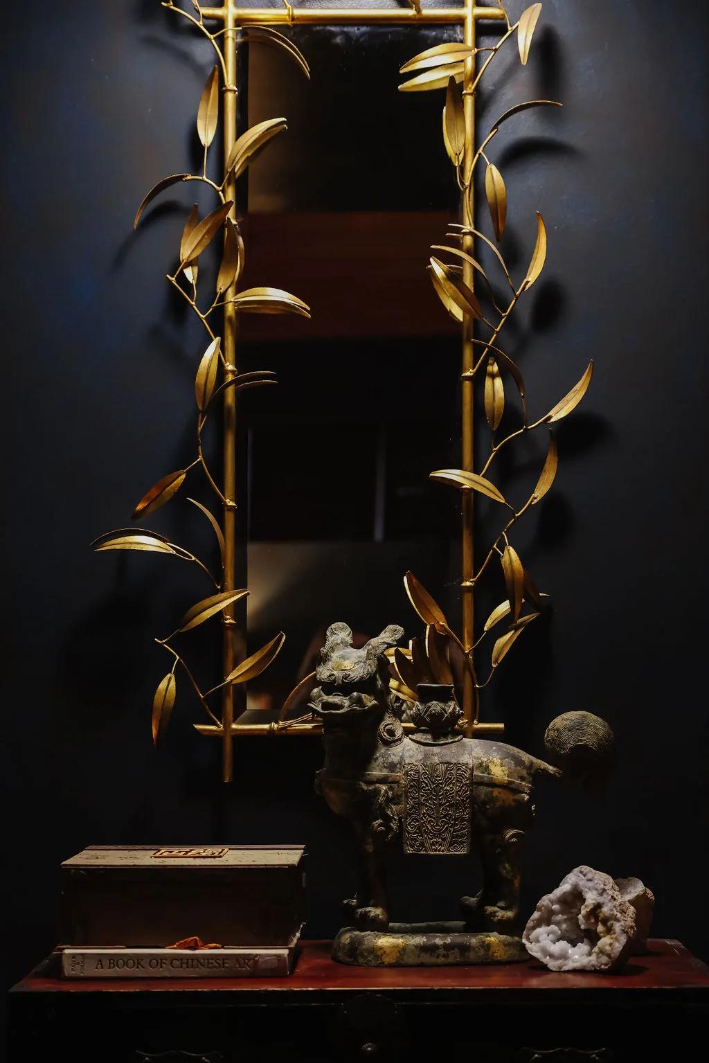 Brass botanical vitrine on a dark wall above a console of stacked books, mineral specimens and a carved figure