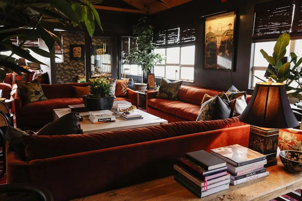 Members lounge with rust velvet sectionals, patterned cushions, tall planting, framed art and a stacked library table
