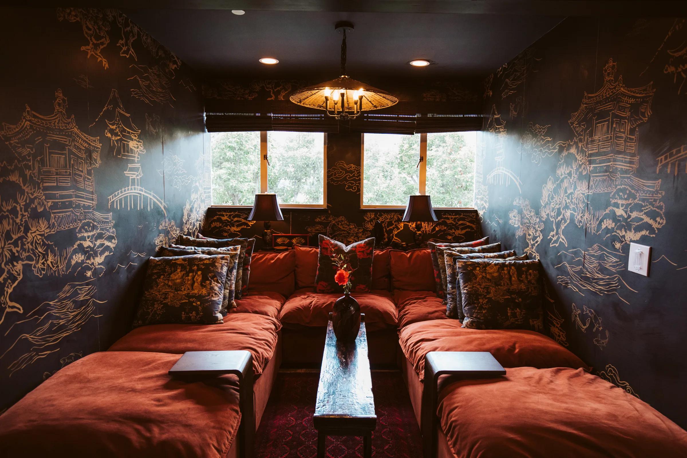 Dark media room with gold chinoiserie wall murals, red velvet chaise seating, twin windows and pendant lamp