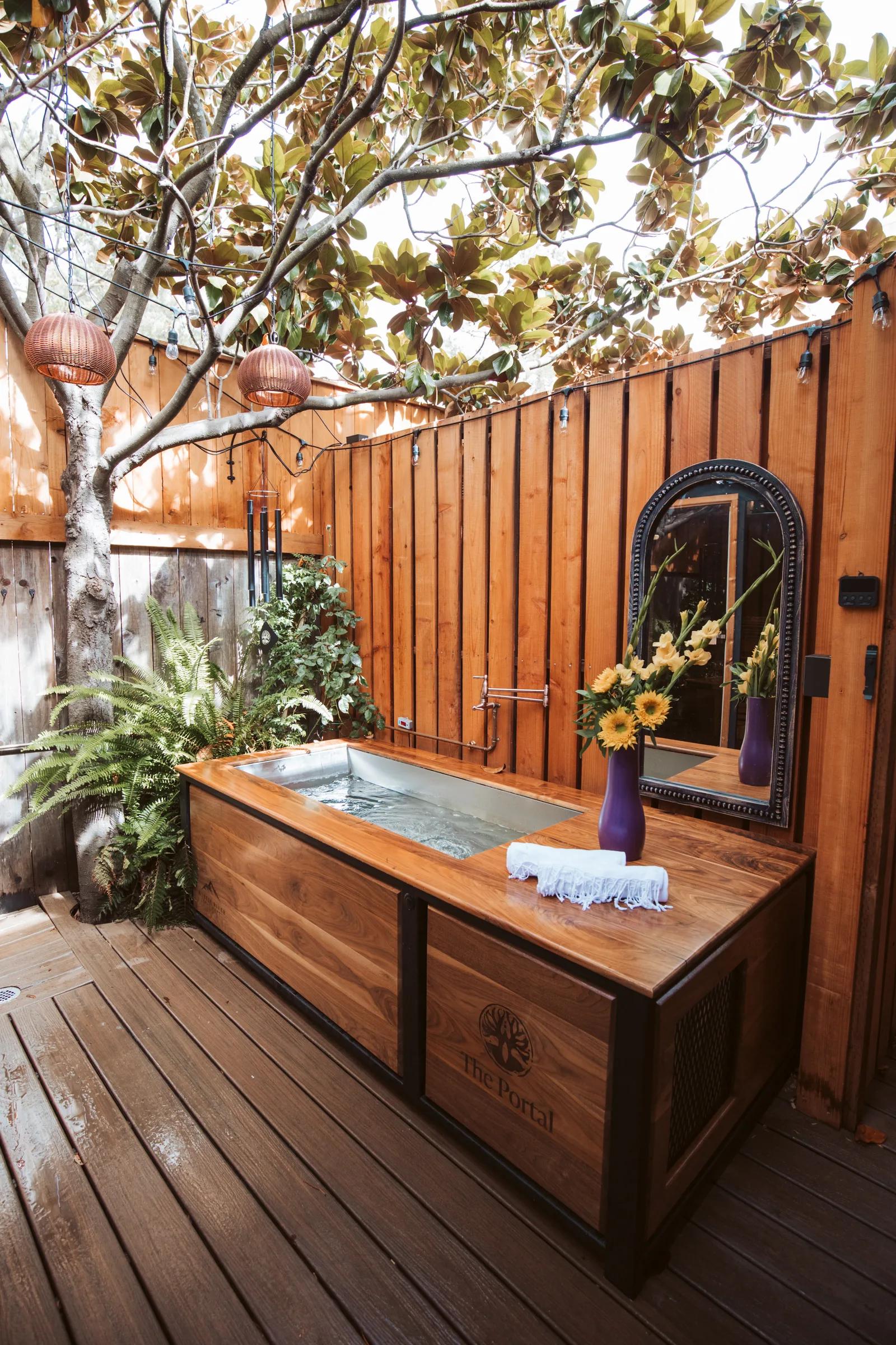 Outdoor walnut cold-plunge tub engraved with The Portal logo, arched mirror, ferns and hanging lanterns