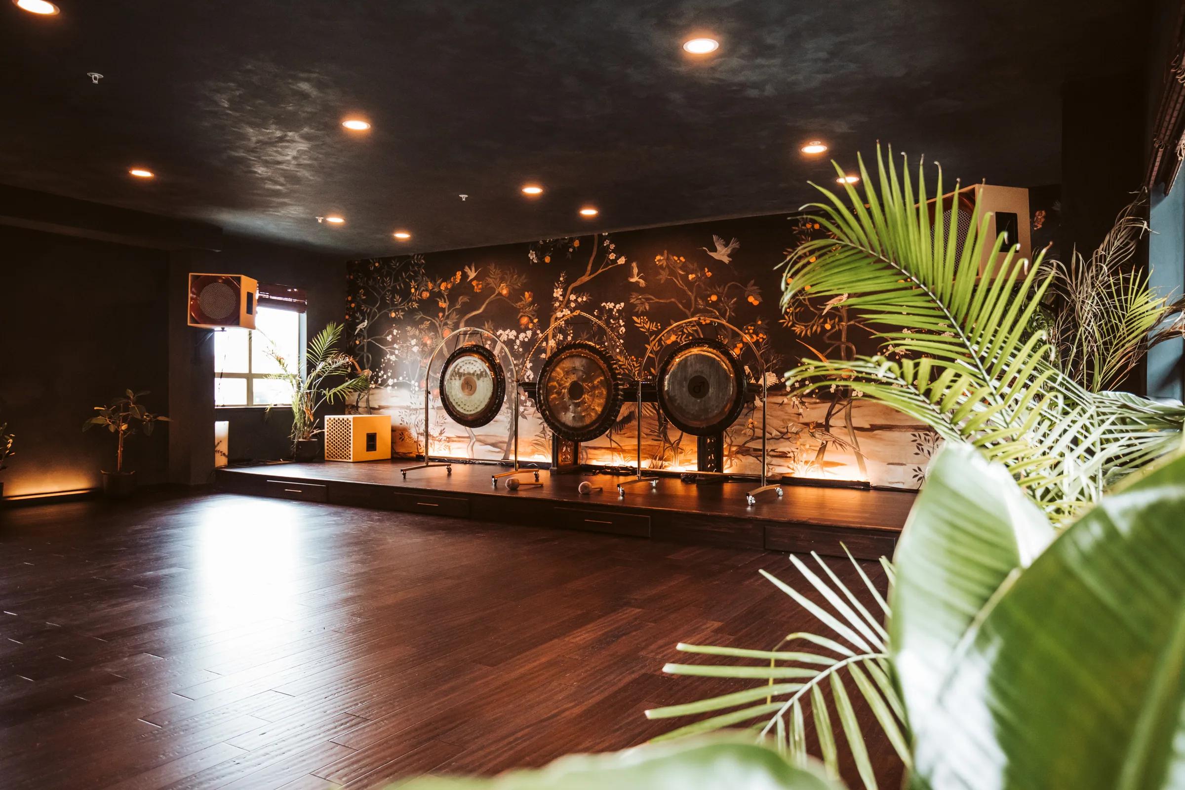 Sound-bath studio with three gongs on a low stage against a lit chinoiserie mural, dark plaster walls and palms