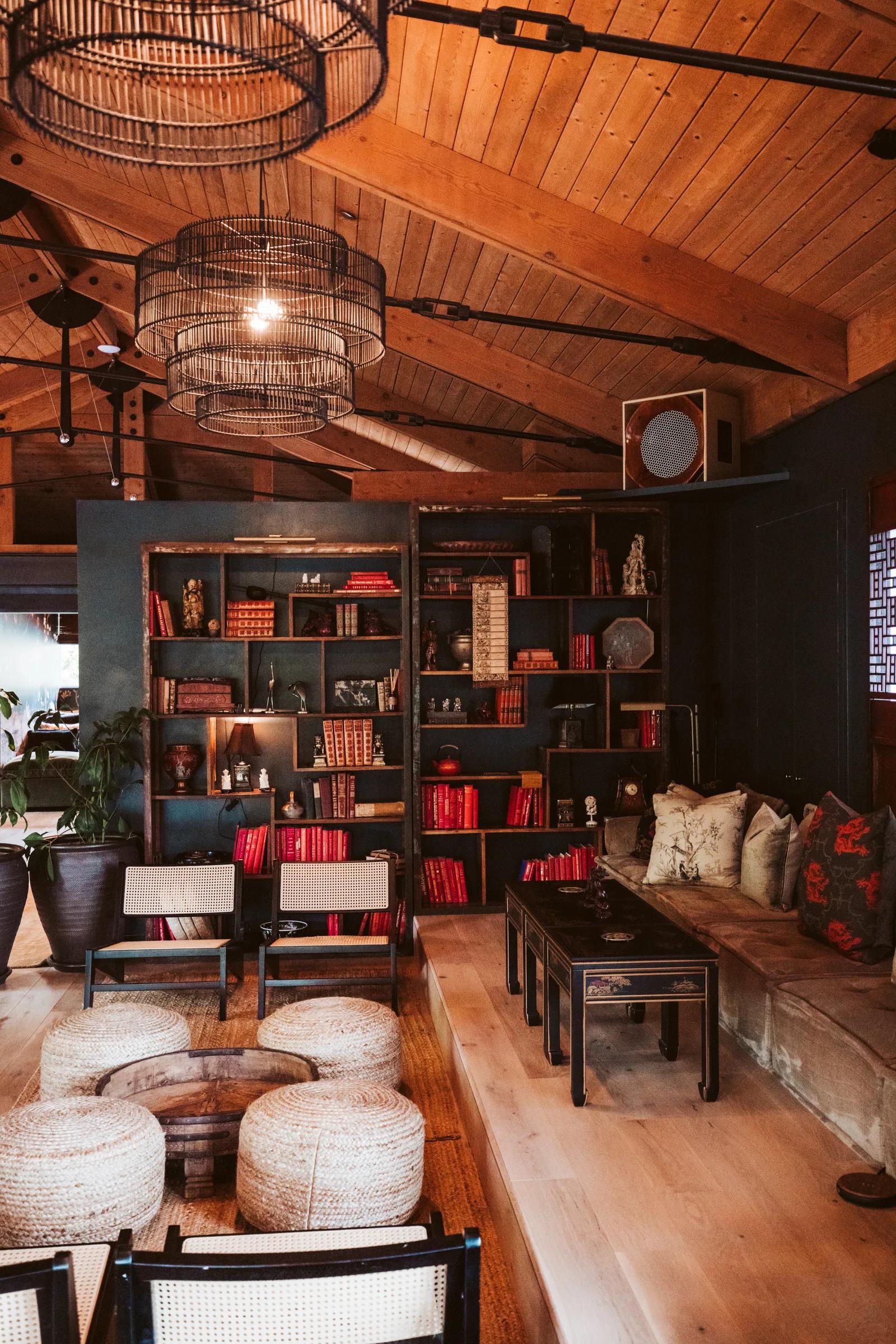 Library corner with dark shelving of red books, cane chairs, woven poufs and a cage pendant under wood ceiling