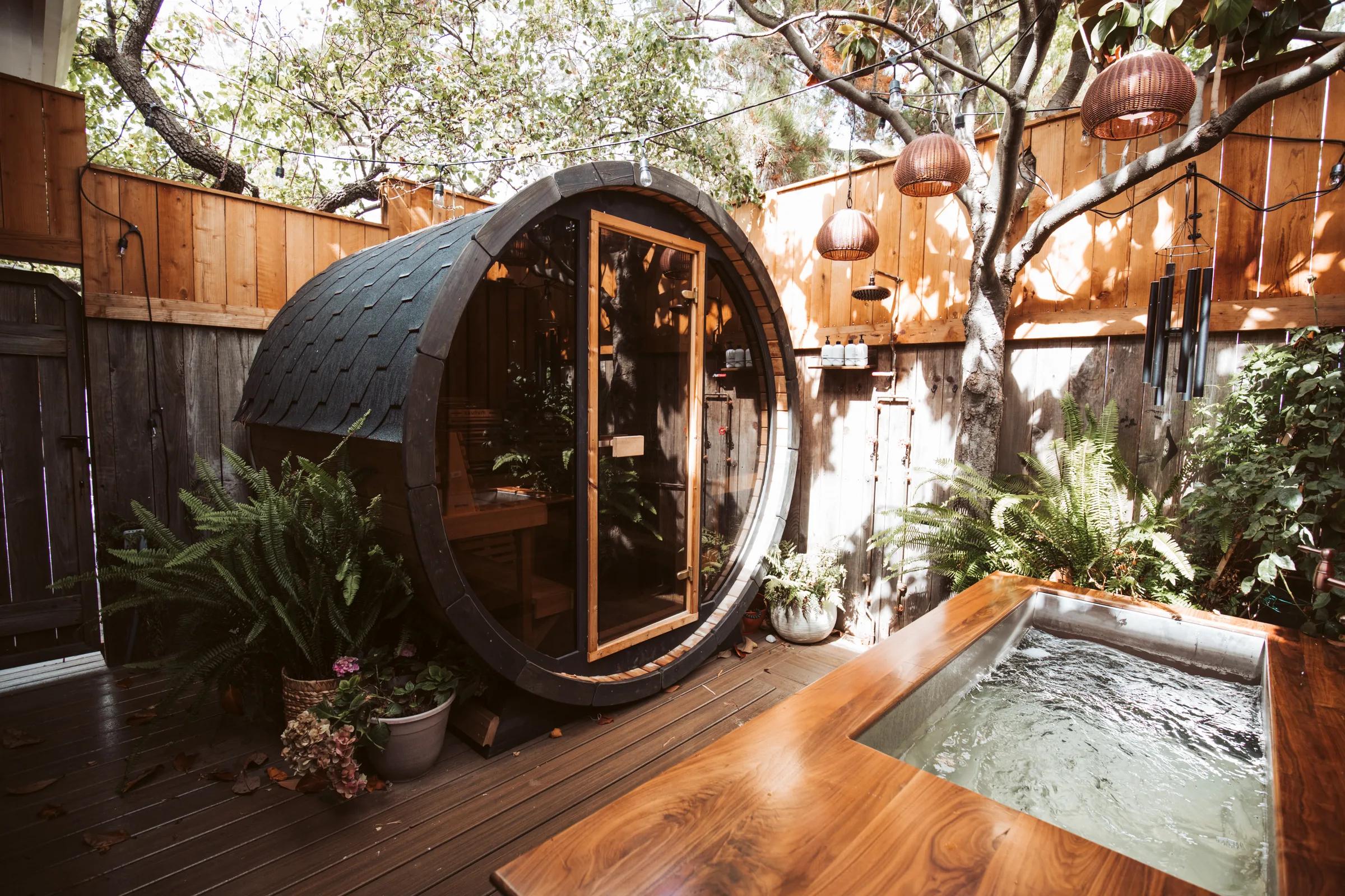 Shingle-roofed barrel sauna beside a walnut cold-plunge tub on a fenced spa deck with ferns