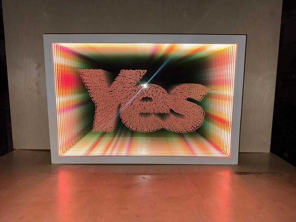 Front view of an infinity-mirror light box with coral Yes lettering over a radiating multicolor LED grid, glow spilling onto a workbench