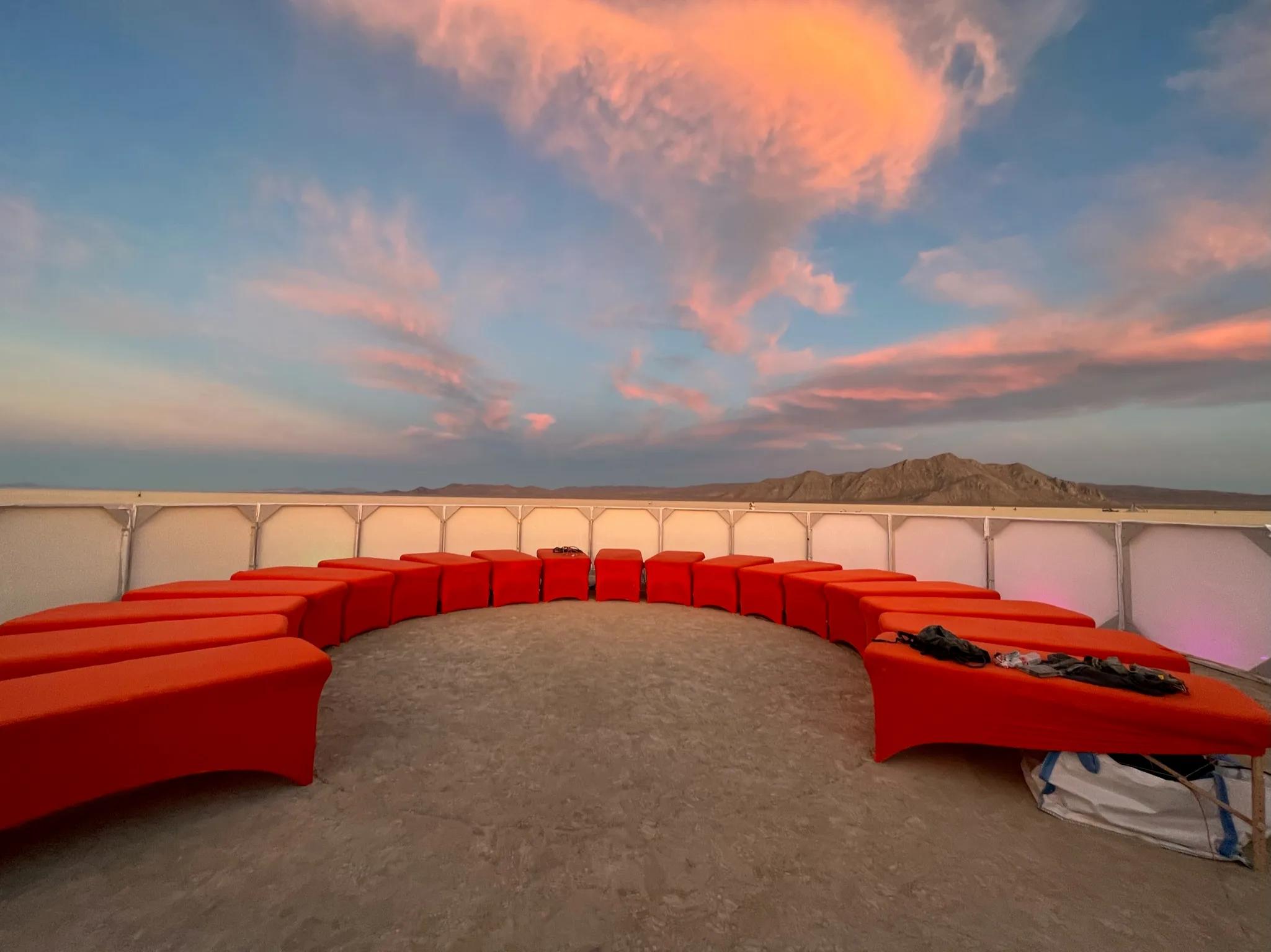 Semicircle of orange upholstered benches inside a white-walled desert enclosure at sunset, mountains beyond