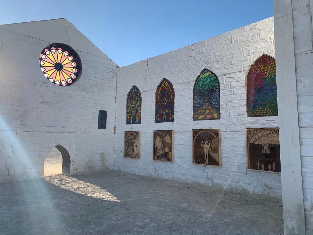 White-painted open-air chapel with rose window, stained-glass arches, and wood-burned panels in desert light