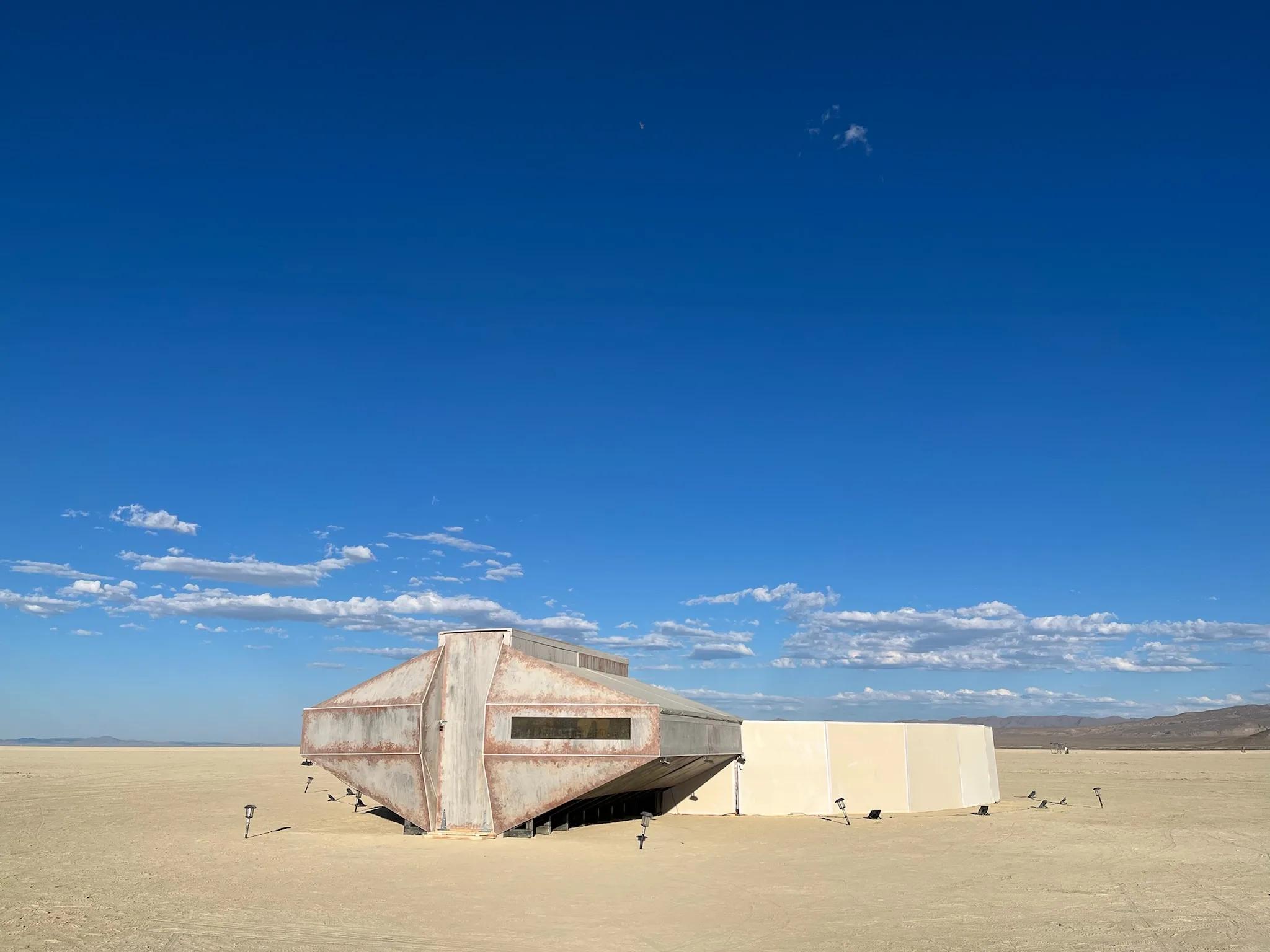 Weathered angular metal pavilion with white fabric wall on open desert playa under deep blue sky