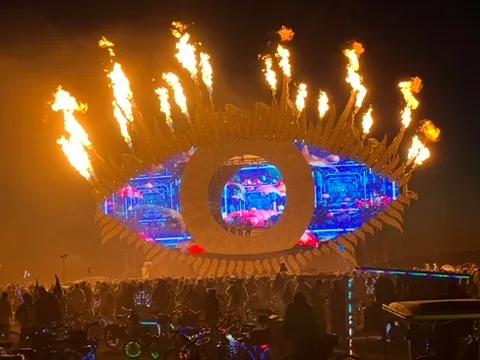Eye-shaped stage firing flame jets from its lashes at night, LED iris screens lit, crowd and bikes in front