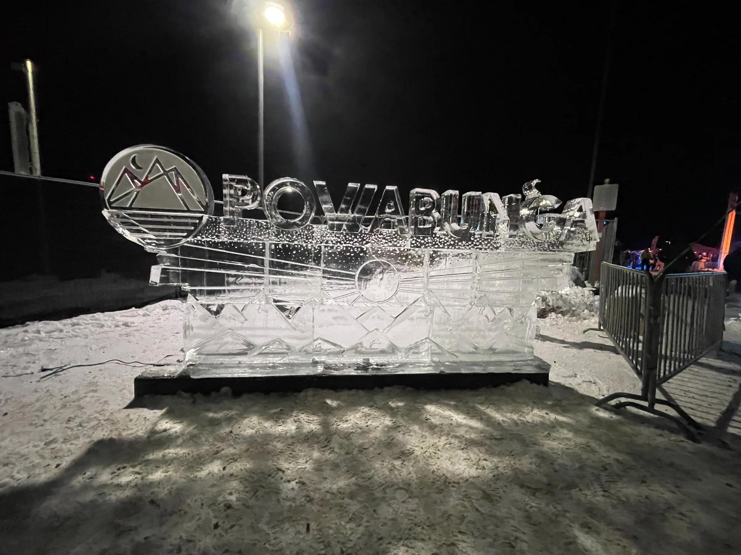Carved ice sculpture reading Powabunga with mountain logo, lit at night on snow beside a barrier fence