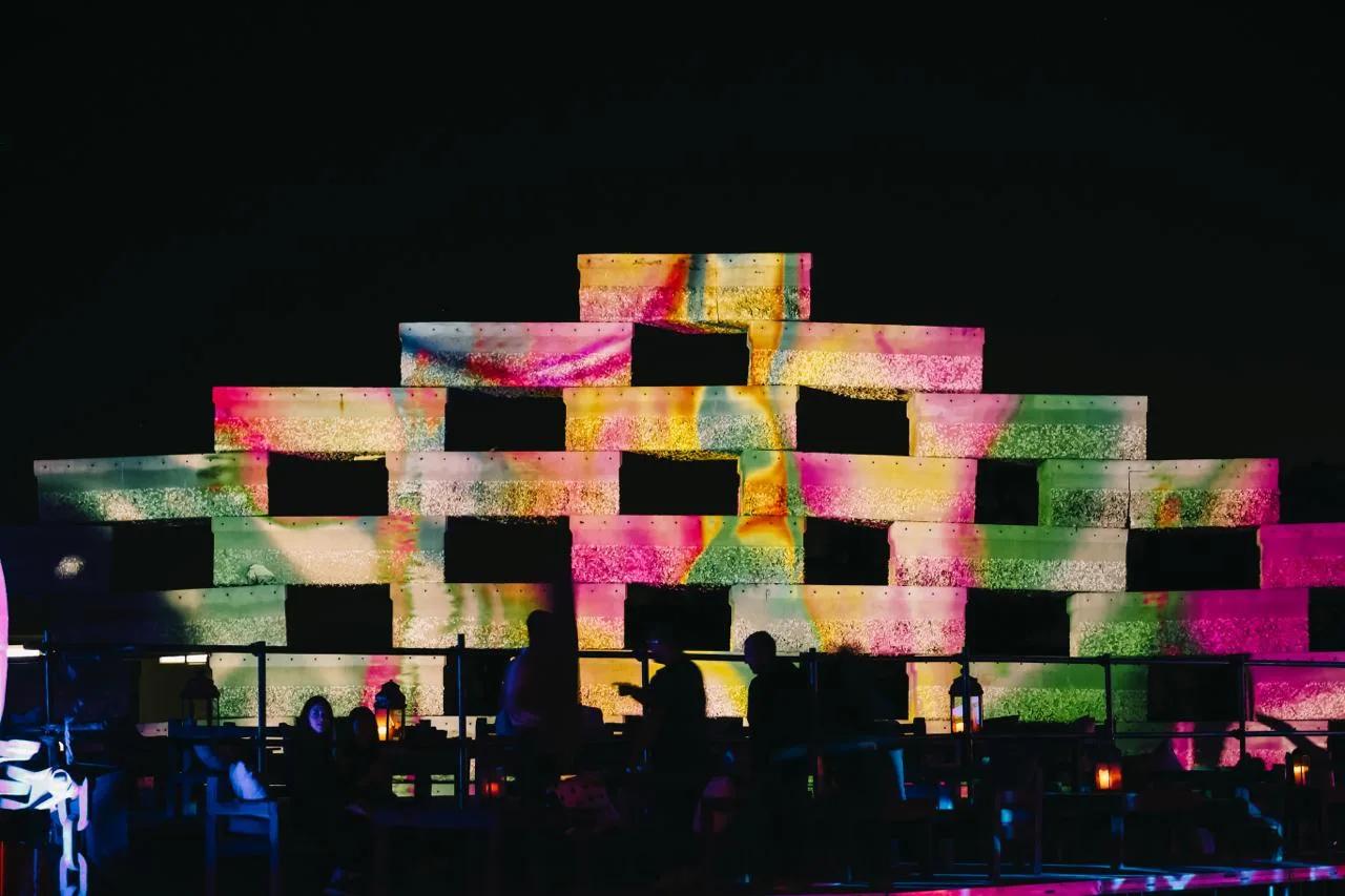 Stacked-block wall with multicolor projection mapping at night, silhouetted guests seated at tables below