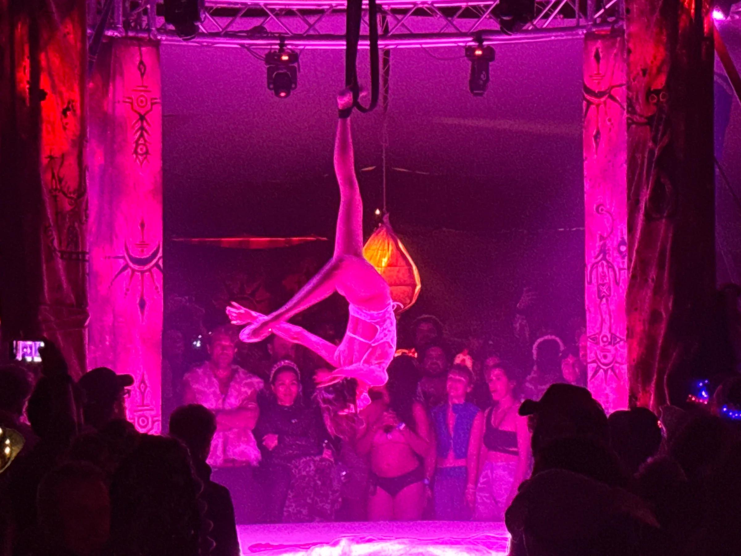 Aerial performer inverted on straps beneath a pink-lit tent rig, painted columns framing the stage
