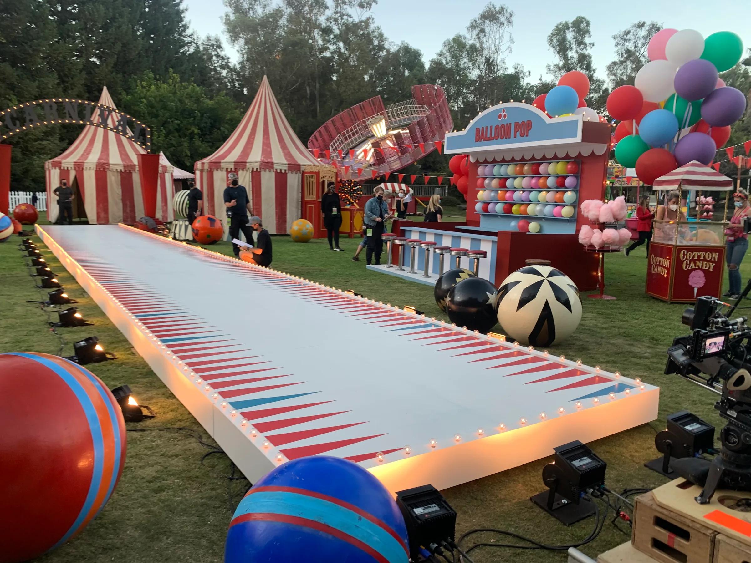Carnival film set with bulb-lit runway, striped circus tents, balloon game booth, and camera rigs at dusk