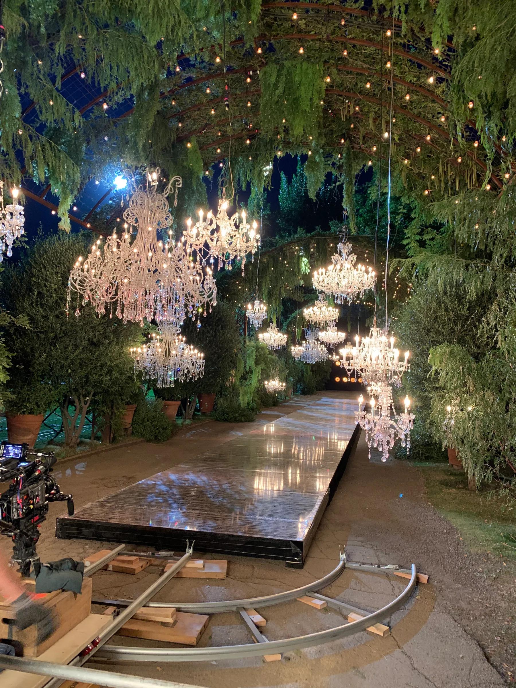 Crystal chandeliers hung from a greenery canopy over a mirrored runway at night, camera dolly track in foreground