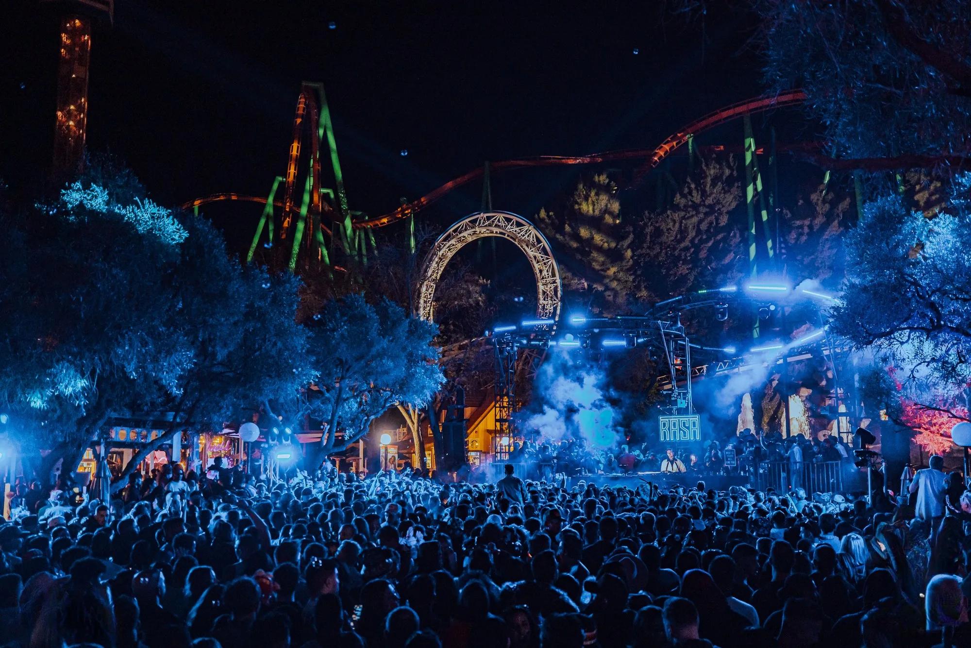 Blue-lit crowd facing a stage beneath an illuminated roller coaster arch and track at night