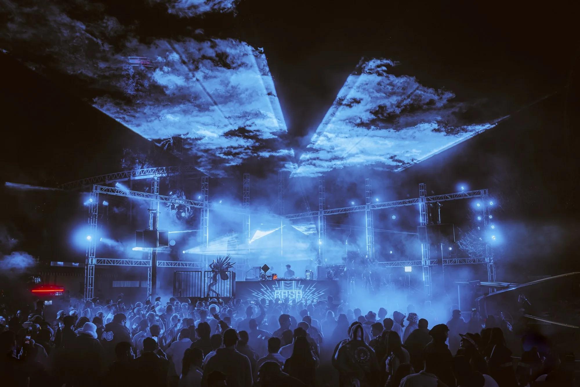 Canopy of blue lasers cutting through haze above a truss-framed RASA main stage and crowd