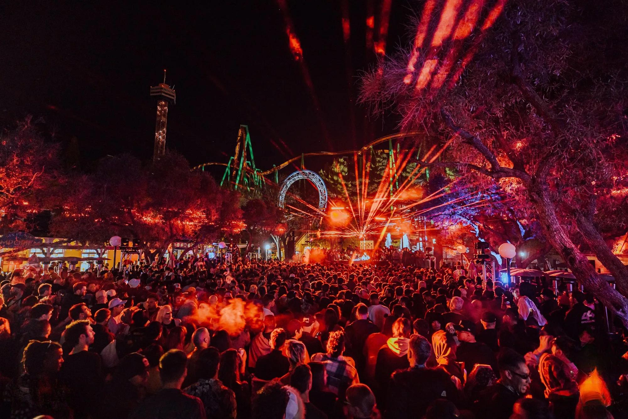 Festival crowd at night under orange lasers, with a lit roller coaster, arch, and drop tower behind the RASA main stage at Six Flags
