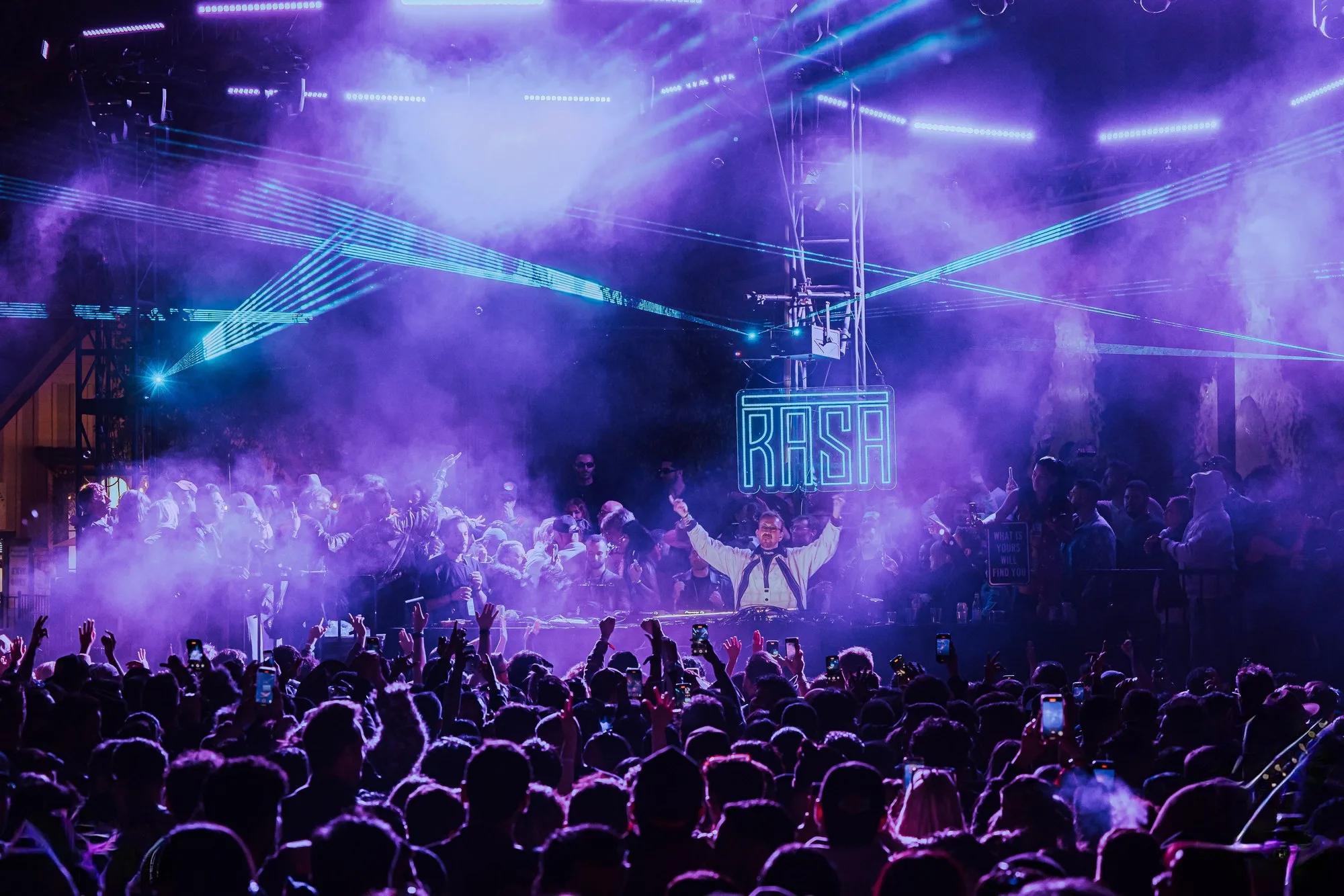 DJ with arms raised beneath a neon RASA sign amid purple haze, lasers, and a phone-lit crowd